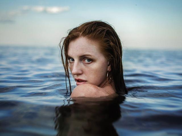 GingerFairys feuchtes, rotes Haar klebt an ihrem Gesicht, während sie mit grünen Augen nach oben blickt, Sommersprossen zeigt und einen ernsten Gesichtsausdruck hat, wobei nur ihre Schultern über dem Wasser sichtbar sind..