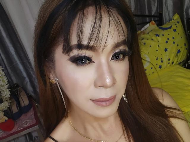 KassandraHotTS has brown hair, dark eyeliner, and long lashes, with a gold nose ring and earrings, wearing a gold necklace, and has a subtle smile, showing her pretty face.