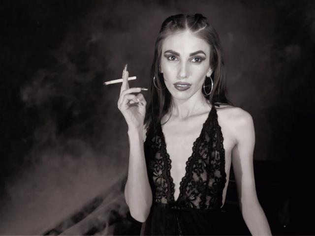 Shares with long brown hair, wearing a black lace dress, holding a cigarette in her right hand, looking at the camera with a serious expression, wearing large hoop earrings.