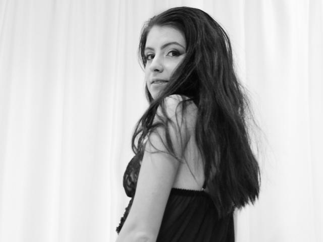 KatherineSicy with long dark hair, wearing a black lace bra, looking over her shoulder, showing her tits, in a black and white photo.