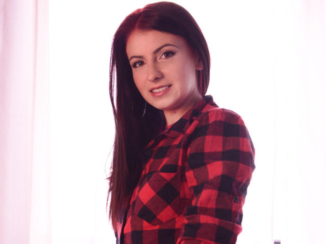 KatherineSicy with long brown hair, wearing a red and black checkered shirt, smiling softly at the camera, showing a cute face and small tits.