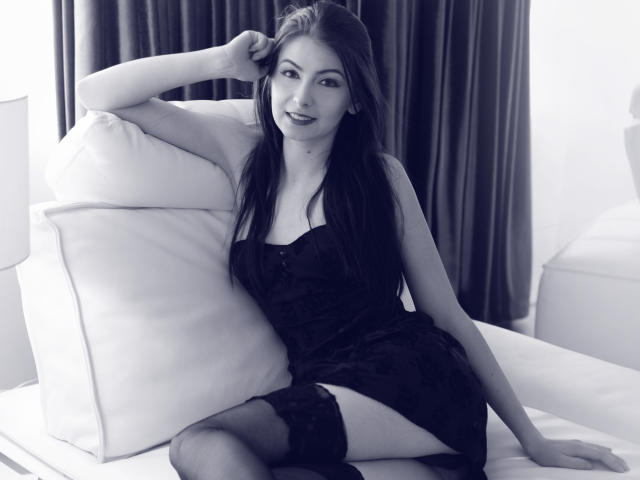 KatherineSicy with long dark hair, wearing a black lace dress, sitting on a white couch, legs apart, showing black thigh-high stockings, and smiling at the camera.