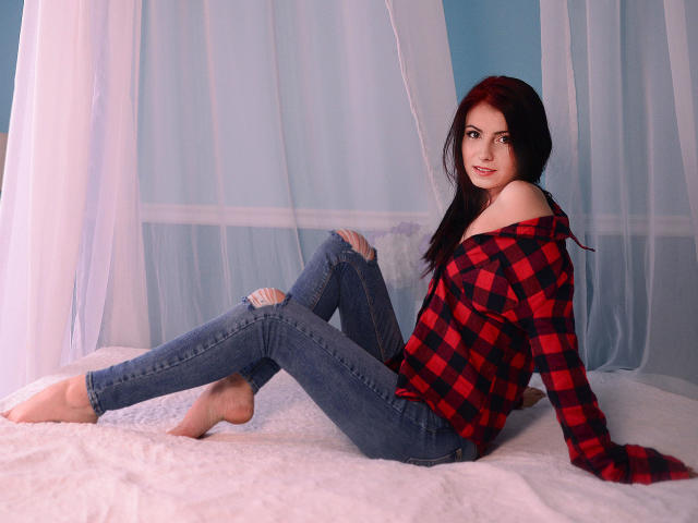 KatherineSicy with red hair, off-shoulder red and black plaid shirt, skinny blue jeans, bare feet, sitting on white bed, looking at camera, sexy, cute.