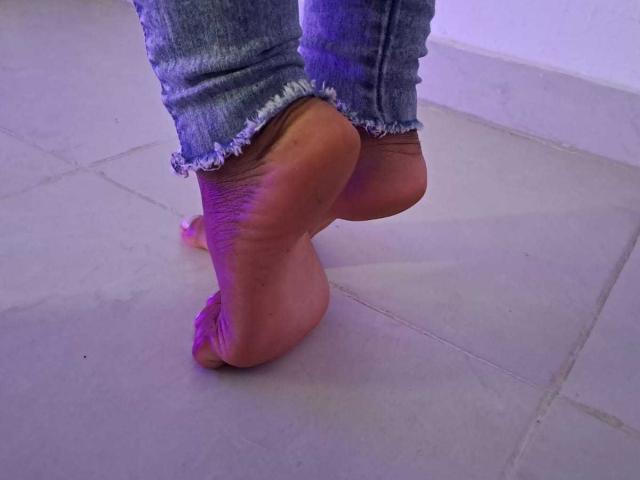 LucyMorga's bare ass and sole of her foot are visible, with her foot raised and showing a slight purple glow, wearing frayed blue jeans.