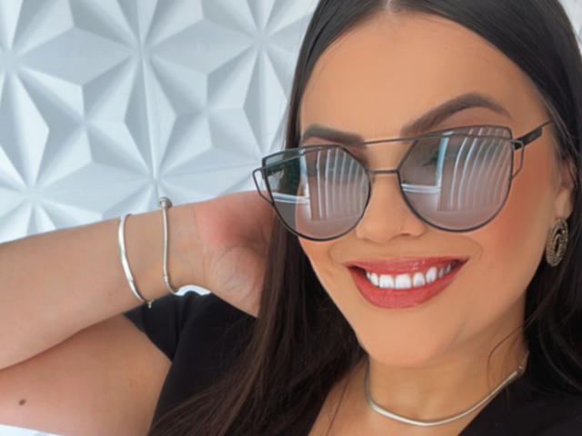 AnnneNaughtyBusty with dark hair, red lipstick, white teeth, black shirt, silver bracelet, and reflective aviator sunglasses, smiling at the camera.
