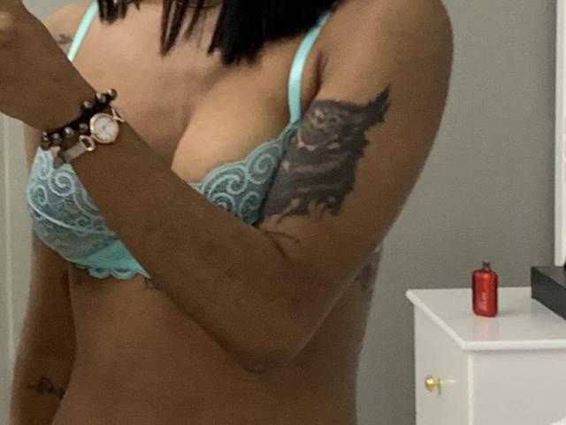 NaomiPortia with dark hair, wearing light blue lace bra and panties, showing tattoos on arm and belly, holding phone, looking down, standing in bedroom with white bed.