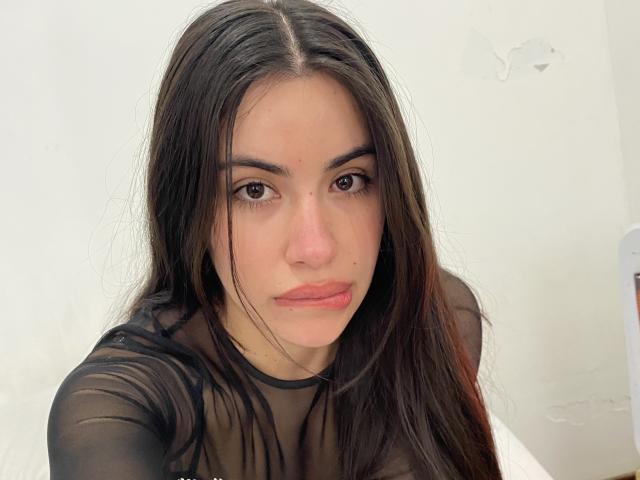 Talita69 has long dark brown hair, brown eyes, and full pink lips. She's wearing a sheer black top, showing her tits. Her expression is serious and slightly pouty.