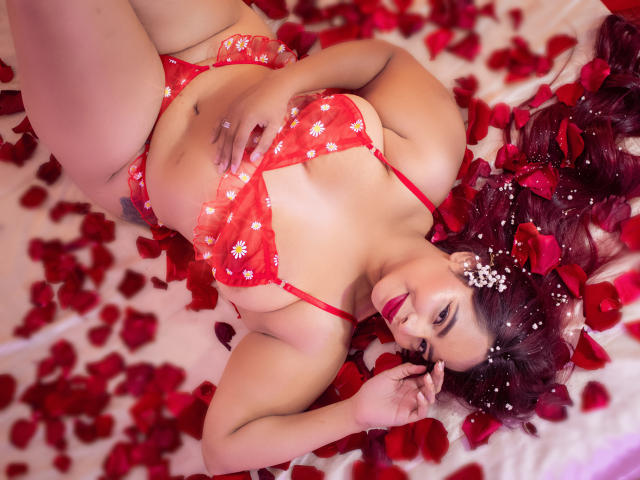 VivianCruzz lies on a bed of red rose petals, wearing a red floral bra and panties, with her hand on her tits and her ass slightly raised.