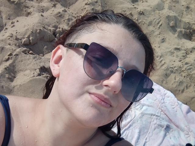 Alifil69 with wet brown hair, wearing dark sunglasses, lying on sandy beach, slight smile, pinkish lips, small freckles on nose, black top, pink and white towel beneath.