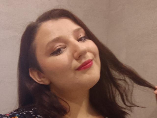 Alifil69 has fair skin, dark brown hair, and red lipstick, smiling slightly with her head tilted. She's holding a lock of hair with her right hand.