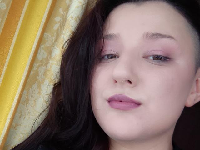 Alifil69 has a pale skin, nose piercing, pinkish eyeshadow, brown eyes, and pink lipstick. Dark brown hair is partially shaved on the left side. She's looking at the camera.