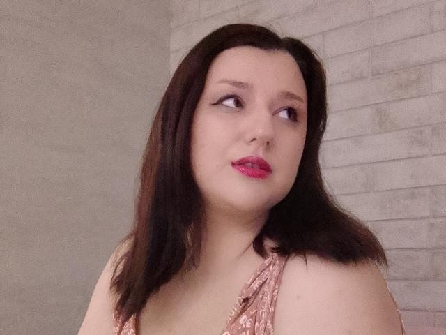 Alifil69 has pale skin, dark brown shoulder-length hair, and pink lipstick. She's wearing a pink patterned top, looking to the side with a subtle smile. Her tits are small.