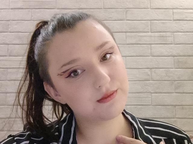 Alifil69 has light skin, brown eyes, and small pink lips. She's wearing a black and white striped shirt with her finger near her chin, and her hair is in a ponytail.