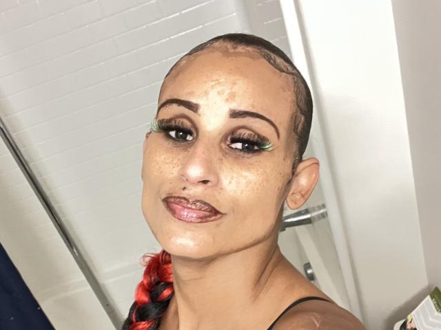 SexyRedBone has a shaved head with braided red and black hair, green eyeliner, and a nose piercing. Skin is shiny with visible freckles and she's smiling at the camera.