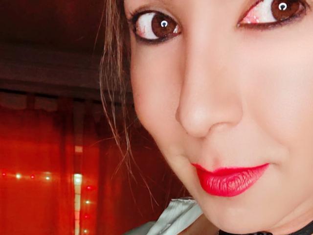 Het gezicht van CamilaMancini, bruine ogen met witte reflecties, rode lippenstift, een lichte glimlach, bruin haar, close-up, haar neus en lippen zijn gecentreerd, rood licht op de achtergrond..