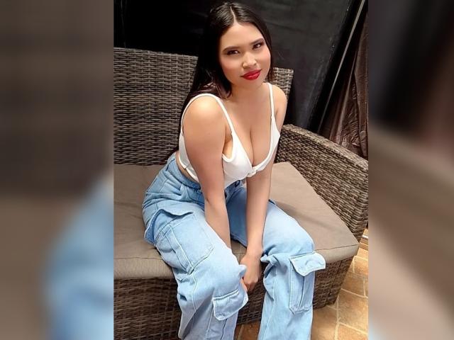 OliviGrace with big tits, wearing a white crop top, blue baggy jeans, and red lipstick, sitting on a wicker bench, looking at the camera with a slight smile.