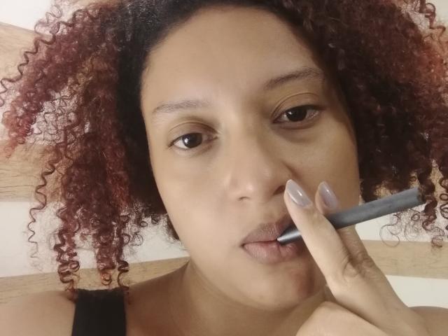 VioletaSwitch with curly red hair, brown eyes, and nude lipstick, holding a blue pencil to her slightly parted lips, showing off her shiny, nude-painted nails.