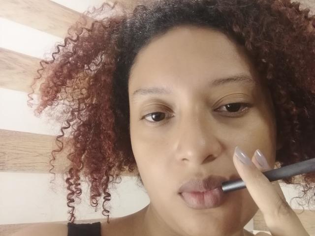 VioletaSwitch with curly red-brown hair, applying greyish lipstick with a black lipstick brush, brown eyes, and nude nail polish, wearing a black top, in a selfie.
