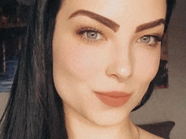 LollaHott has dark brown eyebrows, green eyes, long black hair, and full, matte orange lipstick. Her skin is pale with a slight freckle on her right cheek.