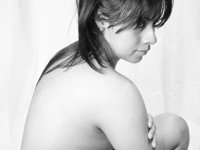 Black-and-white photo of HairyDuchess, topless, with small tits, side profile, holding knee, dark hair, bangs, earring, soft expression, white background, looking downward, gentle light.