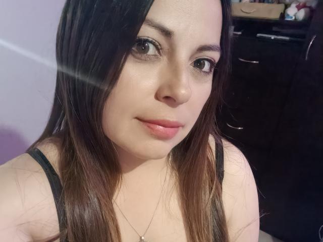 Seher with long brown hair, light skin, and pink lips, wearing a black tank top, has small tits and is looking at the camera with a subtle smile.