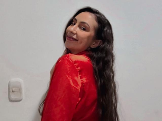 Emma Wells, con cabello largo y oscuro, sonriendo a la cámara, vestida con una blusa roja brillante, mirando por encima del hombro, mostrando su trasero, pared blanca con un interruptor de luz en el fondo..