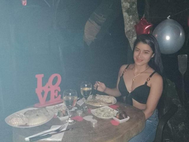 HanaMariana met borsten in een zwarte beha, lang donker haar, lachend, aan een tafel met eten en drinken, en een roze "LIEFDE" bord, terwijl ze een vork vasthouden, in een kamer met weinig licht..