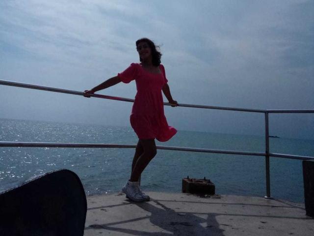 Sarah, con i capelli castano ricci, indossa un vestito rosa e scarpe da ginnastica bianche, appoggiata a una ringhiera di metallo vicino al mare, con il vestito che svolazza al vento..