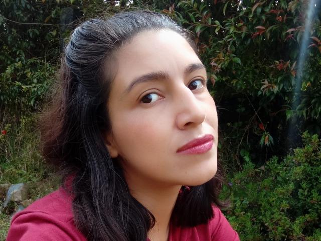 SarahFriendly's face with brown eyes, pink lips, and dark wavy hair, wearing a red shirt, looking slightly to the side in a natural outdoor setting.