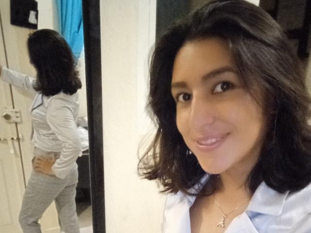 SarahFriendly with wavy black hair, smiling, wearing a white shirt and gray pants, standing with her back to a mirror, showing her ass, in a selfie.