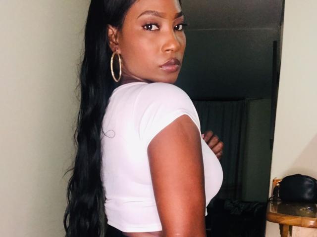 TalianaMoreno with dark skin, long black hair, wearing a white crop top, showing her tits, looking over her shoulder, gold hoop earrings, in a dimly lit room.