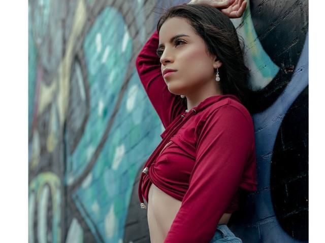 SophyMedina with her tits showing, wearing a red crop top, standing against a graffiti-covered wall, arm raised, looking to the side, with dark hair and earrings.