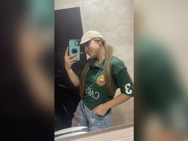 SophyMedina with long brown pigtails, wearing a green shirt with a Texas logo, beige cap, and ripped jeans, taking a mirror selfie in a bathroom. Her phone is teal.