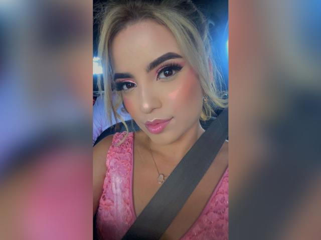 SophyMedina with blonde hair, pink lace dress, and seatbelt, has pink lipstick and dark eyeliner, looking at camera, soft blue and pink blurred background.