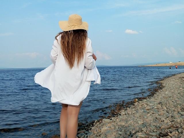 InnaGlory mit langen, braunen Haaren steht an einem felsigen Ufer, trägt ein weißes Kleid und einen Strohhut, blickt auf das blaue Wasser, die Hände an den Seiten. Hintern und Brüste sind nicht sichtbar, der Rücken ist zur Kamera gerichtet..