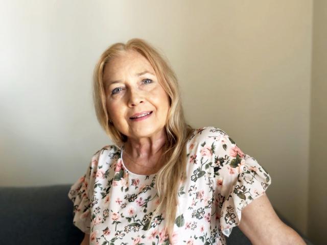 PerfectSofia, blonde woman with light skin, smiling, wearing floral shirt, sitting on gray couch, visible ass, tits, and slight wrinkles, looking up, relaxed, indoors, daytime.