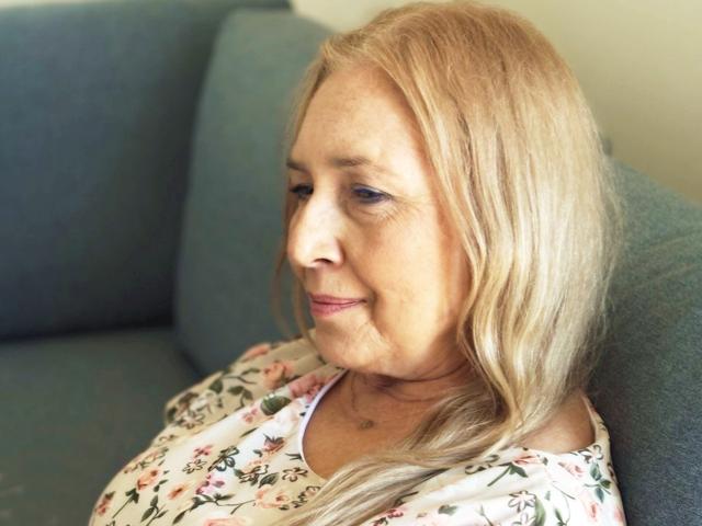 PerfectSofia with blonde hair, slight smile, floral shirt, sitting on blue couch, looking down, visible wrinkles, soft light, gentle expression, slight pink lipstick, calm atmosphere.