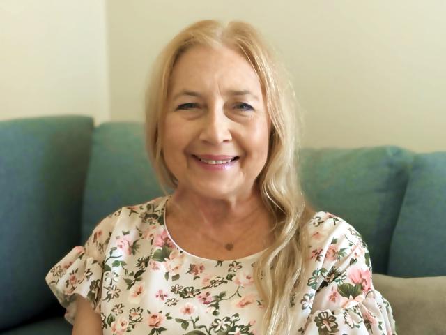 PerfectSofia has blonde hair, smiling, wearing a floral dress with pink and green flowers. Her tits are small, and she's sitting on a green couch.