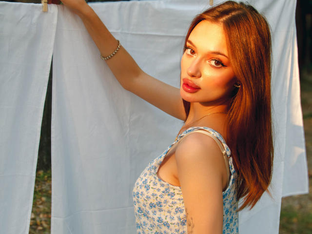 KylieBeaty with red hair, blue floral dress, holding white clothes, looking over shoulder, sunlight on face, bracelet on wrist, soft smile, cute, sexy, upper body, daytime.
