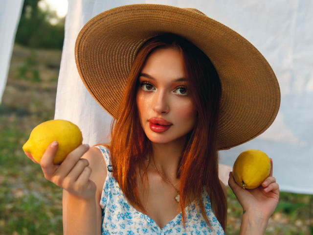 KylieBeaty with red hair, wearing a big straw hat, holding two lemons, in a blue floral dress, looking at the camera with a slight smile. Boobs visible under dress.