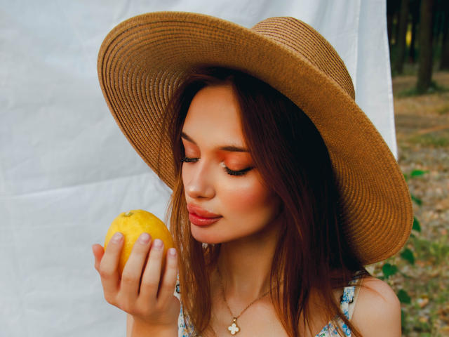 KylieBeaty with brown hat, holding yellow lemon, eyes closed, wearing floral dress, red lipstick, and necklace, standing outdoors, soft sunlight, gentle smile, subtle makeup, pretty, cute.