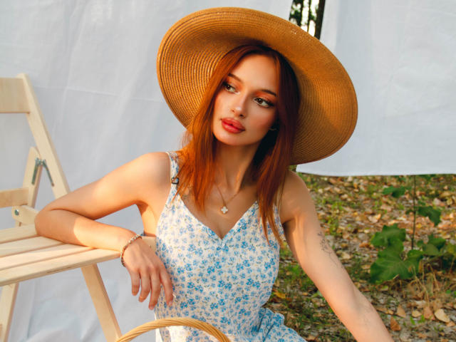 KylieBeaty with red hair, wearing a straw hat, blue floral dress, and red lipstick, sitting on a wooden chair, showing her tits and a tattoo on her left arm.