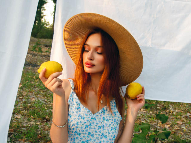 KylieBeaty with red hair, wearing a straw hat, blue floral dress, holding two yellow lemons, bracelet on right arm, eyes closed, sunlit, green grass background.