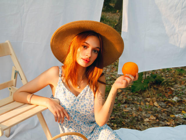 KylieBeaty with orange hair, wearing a sunhat, floral dress, holding an orange, sitting on a wooden chair, sunlight, red lipstick, tattoo on right forearm, white fabric backdrop.