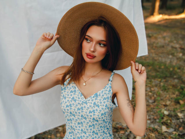 KylieBeaty with brown hat, blue floral dress, red lipstick, holding brim, showing tattoos on left arm, silver bracelet, and necklace, standing outdoors.