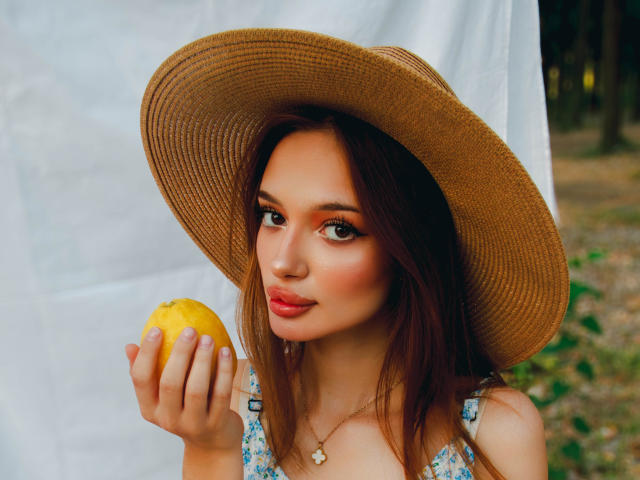 KylieBeaty with a big brown hat holding a yellow lemon, smiling with pink lips, brown eyes, and wearing a floral dress, standing outdoors, with a delicate necklace.
