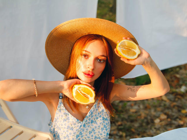 KylieBeaty with red hair, wearing a sun hat, holding orange halves, in a blue floral dress, has a tattoo on her right arm, and a bracelet on her left wrist.