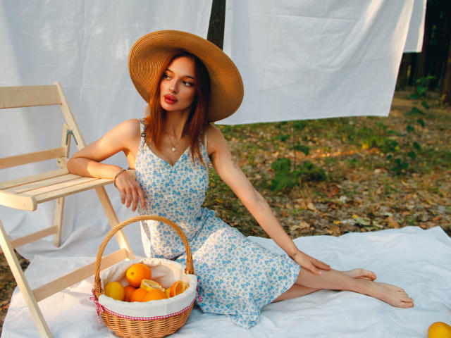KylieBeaty, wearing a blue floral dress and straw hat, sits on a white cloth with a wicker basket of oranges. She's barefoot, leaning on a wooden chair.