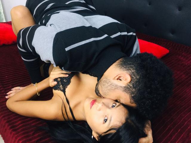 Karen e Alex, com pele escura e cabelo cacheado, estão em uma cama vermelha, beijando o peito de uma mulher que usa um sutiã de renda preta, tem lábios vermelhos e cabelo preto longo..