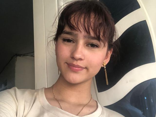 Melody Mansson tem cabelo castanho, pele de tom médio, nariz pequeno e está sorrindo com lábios rosados. Ela veste uma camisa branca, brincos de ouro e um colar..